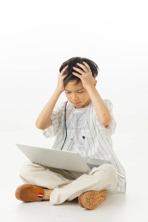 Téléchargez les photos : Heureux petit garçon asiatique assis en tapant ordinateur portable tandis que confus isolé sur fond blanc, concept d'apprentissage en ligne, En regardant la caméra et la composition complète du corps. - en image libre de droit