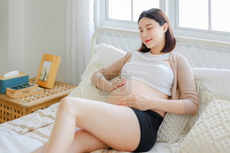 Téléchargez les photos : Jeune asiatique heureuse femme enceinte est assis et relaxant sur le lit et toucher son ventre. Plan familial, grossesse, maternité, personnes et concept d'attente - en image libre de droit