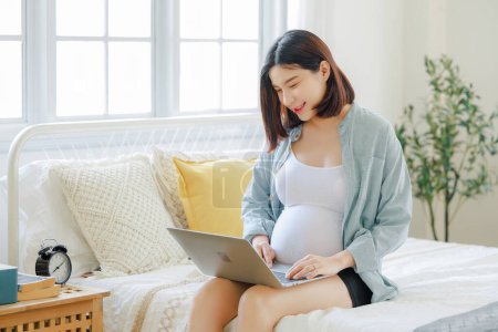 Téléchargez les photos : Jeune asiatique heureuse femme enceinte est assis et relaxant sur le lit et toucher son ventre. Plan familial, grossesse, maternité, personnes et concept d'attente - en image libre de droit