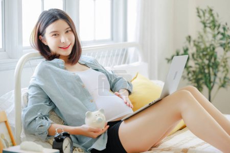 Foto de Joven asiática feliz embarazada está sentada y relajándose en la cama y tocando su vientre. Plan familiar, embarazo, maternidad, personas y concepto de expectativa - Imagen libre de derechos