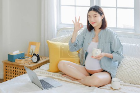 Téléchargez les photos : Jeune asiatique heureuse femme enceinte est assis et en utilisant ordinateur portable tenant tirelire montrer OK signe sur le lit et toucher son ventre. Plan familial, grossesse, maternité, personnes et concept d'attente - en image libre de droit