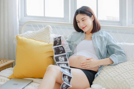 Photo for Young Asian happy pregnant woman is and holding ultrasound 4d scan image touching her belly. Family plan, pregnancy, motherhood, people and expectation, child and Maternity prenatal care concept - Royalty Free Image