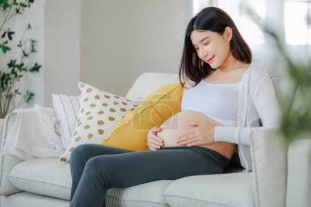 Photo for Young Asian happy pregnant woman is sitting on sofa and touching her belly. Family plan, pregnancy, motherhood, people and expectation, Child and Maternity prenatal care concept. - Royalty Free Image
