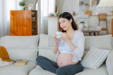 Téléchargez les photos : Femme enceinte buvant de l'eau ou du café assis sur le canapé. Jeune femme enceinte se reposer à la maison avec du verre, copier l'espace. Grossesse, santé, concept de soif. - en image libre de droit