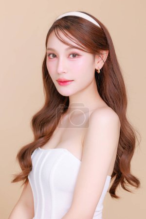 Téléchargez les photos : Jolie beauté asiatique femme cheveux longs bouclés avec maquillage coréen visage éclatant et visage sain portrait de la peau sourire sur fond beige isolé. Cosmétologie, Chirurgie plastique. - en image libre de droit