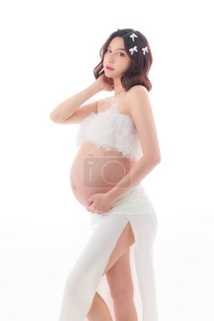 Téléchargez les photos : Joyeux jeune asiatique femme enceinte avec le style de maquillage coréen sur le visage et la peau parfaite toucher ventre sur fond blanc isolé. Concept de grossesse, Soins prénatals de maternité. - en image libre de droit