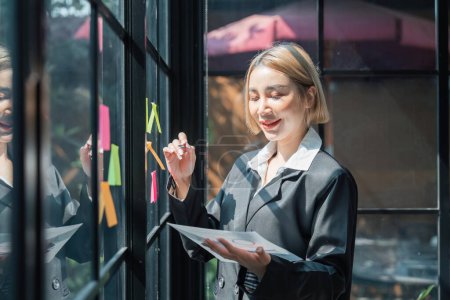Téléchargez les photos : Fille asiatique femme entrepreneur de petite entreprise mettre un adhésif collant notes dans le mur de verre dans le bureau lors de l'analyse de la formulation de stratégies d'affaires. - en image libre de droit