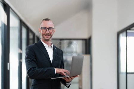 Foto de Hombre de negocios guapo sonriendo usando el ordenador portátil mientras está de pie en la oficina. - Imagen libre de derechos