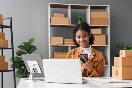Foto de Sme idea de negocios. Mujer afroamericana vendiendo en línea en casa con computadora portátil para tomar pedidos y entregar cajas amarillas ideas de entrega en línea. - Imagen libre de derechos