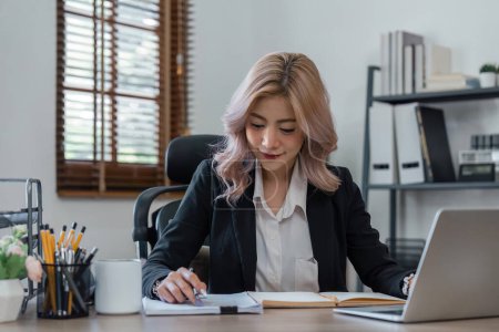 Téléchargez les photos : Portrait d'une femme d'affaires souriante assise au bureau, utilisant un ordinateur portable et écrivant dans un tutoriel pour ordinateur portable. - en image libre de droit