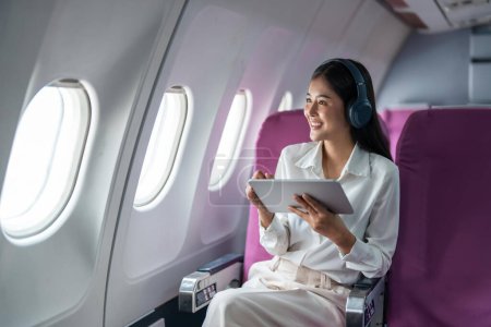 Foto de Joven mujer de negocios asiática o pasajera con auriculares inalámbricos y trabajando con la tableta durante el vuelo. - Imagen libre de derechos