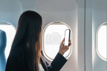 Foto de Una pasajera se sienta en el asiento de la ventana en clase económica, usando su teléfono inteligente, sosteniendo una maqueta de pantalla blanca del teléfono móvil. imagen de primer plano. - Imagen libre de derechos