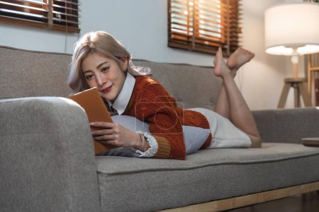 Foto de Joven mujer asiática relajarse en el sofá en casa acogedora leer libro interesante. Descanso femenino feliz en el sofá en la sala de estar disfrutar de novela o historia en fin de semana. - Imagen libre de derechos