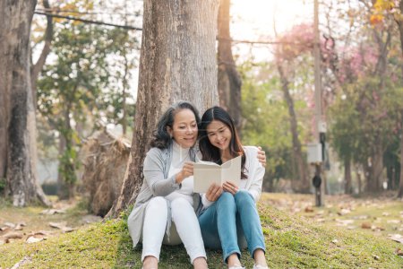 Téléchargez les photos : Belle petite-fille et grand-mère assis sous l'arbre dans le parc vert, lire un livre ensemble. - en image libre de droit