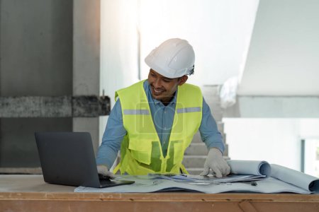 Photo for Asian male engineer working with drawings inspection on laptop on construction site at work desk in office. - Royalty Free Image