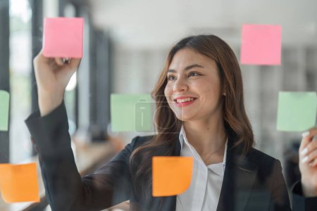 Photo for Close up of focused business female write down on colorful sticky notes manage list, concentrated business woman work on startup brainstorm collaborate plan on stickers on glass wall. - Royalty Free Image