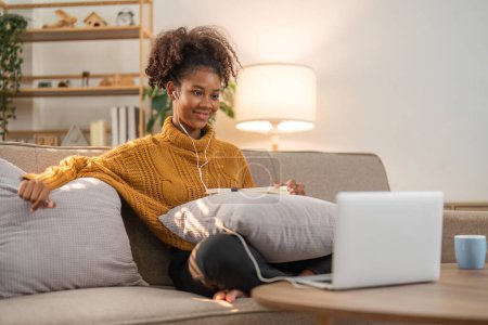 Téléchargez les photos : Image de femme afro-américaine joyeuse utilisant un ordinateur portable et écouter de la musique tout en étant assis sur une chaise dans le salon. - en image libre de droit