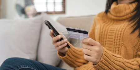 Photo for Young Asian woman sitting on sofa in reception room at home is paying with credit card using mobile phone. Online payment, online shopping concept. - Royalty Free Image