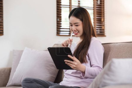 Foto de Hermosa atractiva joven asiática sentada en un sofá gris y usando una tableta digital en la sala de estar por la mañana. - Imagen libre de derechos