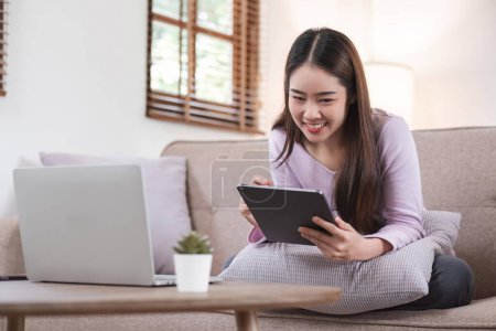 Téléchargez les photos : Belle jeune femme asiatique attrayante assise sur un canapé gris et utilisant une tablette numérique dans le salon le matin. - en image libre de droit