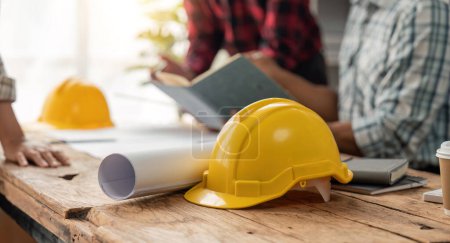 Yellow hard hat on table and house design print design with construction team planning start new project plan in office at construction site partner and contractor.