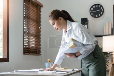 Foto de Joven mujer asiática de negocios sosteniendo archivo de documento y mirando el documento en la oficina del lugar de trabajo, pila de papel de sobrecarga de negocios. - Imagen libre de derechos