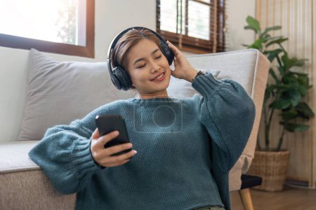 Photo for Woman listening to music, using headphones, sitting on couch in living room, Happy young lady with closed eyes enjoying music. - Royalty Free Image