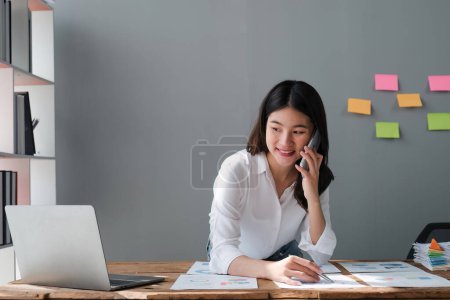 Téléchargez les photos : Heureuse jeune femme asiatique parlant sur le téléphone mobile et souriant tout en étant assise à son lieu de travail au bureau. - en image libre de droit