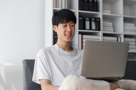 Téléchargez les photos : Jeune homme souriant navigue à son ordinateur portable, assis à la maison sur le canapé confortable à la maison, portant une tenue décontractée. - en image libre de droit