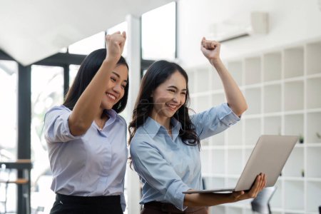 Foto de Dos jóvenes empresarias muestran alegre expresión de éxito en el trabajo sonriendo felizmente con una computadora portátil en una oficina moderna. - Imagen libre de derechos