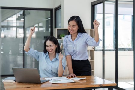 Foto de Dos jóvenes empresarias muestran alegre expresión de éxito en el trabajo sonriendo felizmente con una computadora portátil en una oficina moderna. - Imagen libre de derechos