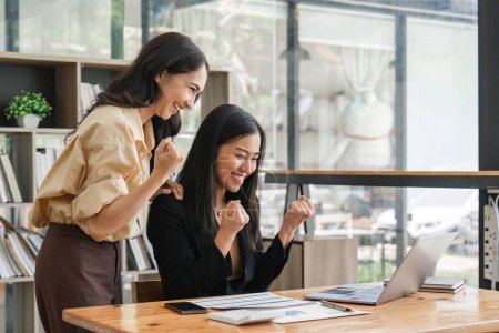 Téléchargez les photos : Deux jeunes femmes d'affaires expriment joyeusement leur succès au travail en souriant joyeusement avec un ordinateur portable dans un bureau moderne. - en image libre de droit