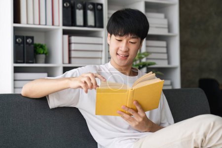 Photo for Portrait of young man holding paper book or diary, sitting on couch at home in living room, reading literature or checking his working schedule plan. - Royalty Free Image