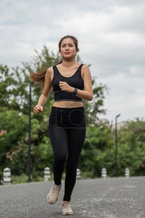 Téléchargez les photos : Femme heureuse portant des vêtements de sport faisant du jogging dans le parc. Jeune belle asiatique femelle en soutien-gorge de sport courir en plein air. Exercice d'entraînement le matin. Concept de mode de vie sain et actif. - en image libre de droit