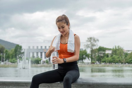 Foto de Asiática hermosa mujer corredor beber agua de una botella. Joven bonita hembra descansando después de hacer ejercicio en el parque. - Imagen libre de derechos