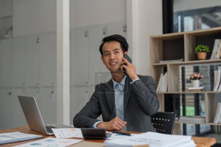 Téléchargez les photos : Homme d'affaires positif parlant sur téléphone portable, assis sur le lieu de travail avec ordinateur portable dans le bureau moderne. - en image libre de droit
