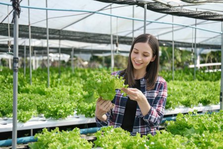Photo for Young farmer female smiling and holding mobile smart tablet with hydroponic fresh green vegetables produce in greenhouse garden nursery farm, smart farming, agriculture business concept. - Royalty Free Image