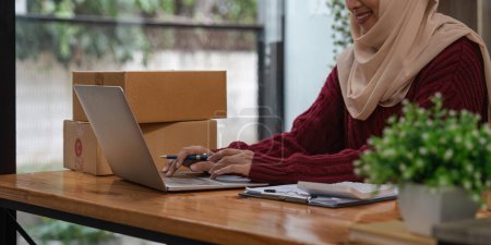 Téléchargez les photos : Musulmane asiatique femme freelance sme entreprise achats en ligne travail et check-list ordre avec boîte en carton sur la table à la maison - Business expédition en ligne et concept de livraison
. - en image libre de droit