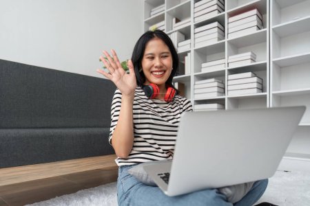 Portrait of happy attractive asian woman and having video call, waving hand at laptop, having online meeting or conversation while relaxing at home.