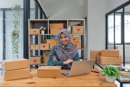 Téléchargez les photos : Entreprise souriant femme musulmane propriétaire préparant le produit pour le client. petit magasin de commerce électronique en ligne. - en image libre de droit