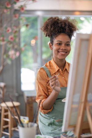 Téléchargez les photos : Une Afro-Américaine qui aime l'art ou une artiste féminine tient une palette de peinture et des pinceaux tout en peignant joyeusement sur papier à l'atelier du studio. - en image libre de droit