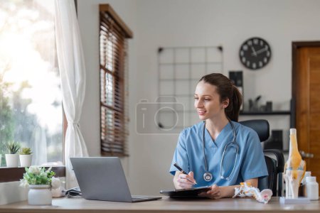 Foto de Retrato de alegre sonriente joven médico en uniforme médico azul escribiendo en el ordenador portátil, sentado en el escritorio cerca de la ventana en la oficina moderna de la clínica médica. - Imagen libre de derechos