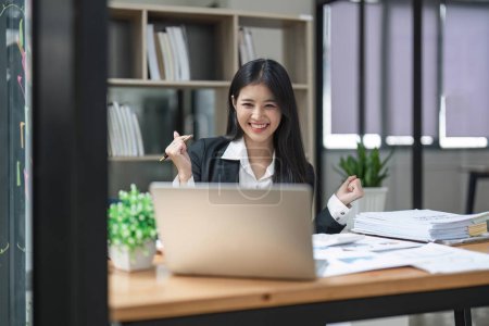 Foto de Feliz joven mujer de negocios asiática celebrando el éxito en el trabajo en la oficina mirando el ordenador portátil entusiasmado con el crecimiento del comercio financiero, metas logro buenos resultados en línea. - Imagen libre de derechos