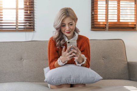 Photo for Happy relaxed young woman asian sitting on couch using cell phone, smiling lady asian holding smartphone, looking at cellphone enjoying doing online ecommerce shopping in mobile apps. - Royalty Free Image