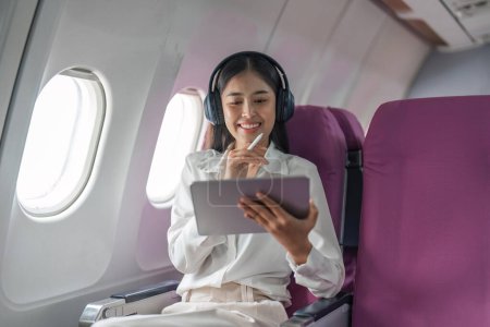 Foto de Avión de pasajeros mujer de negocios que trabaja en cabina de avión durante el vuelo con en vuelo wifi escribir escritura en la tableta de ordenador escuchando música con auriculares. - Imagen libre de derechos