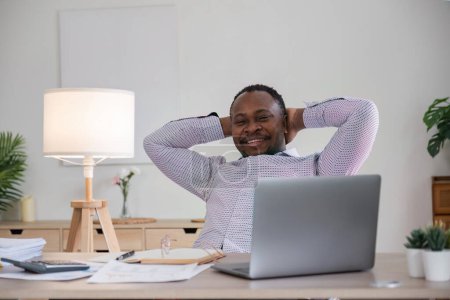 Photo for Smiling black man using laptop at home in living room. Happy mature businessman send email and working at home. African american man typing on computer with paperworks and documents on table. - Royalty Free Image