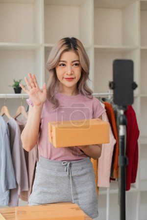 Photo for Woman entrepreneur showing clothes product on live online social media streaming and packing into cardboard box to preparing for shipping customer delivery at home. - Royalty Free Image