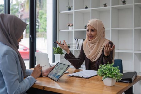 Téléchargez les photos : Jeune femme musulmane d'affaires discutant avec des amis assis ensemble, travaillant avec une tablette et des documents financiers au bureau. - en image libre de droit