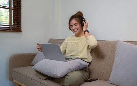 Foto de Mujer joven en los auriculares usando el ordenador portátil escuchando música relajarse sentado en el sofá en casa. - Imagen libre de derechos