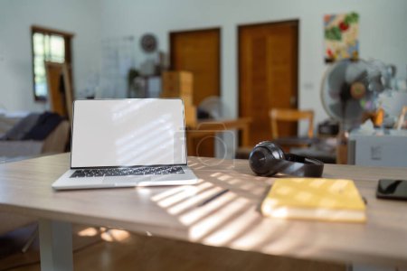Photo for White screen of laptop, headphones and notebook on the wood table with blurred background of home. - Royalty Free Image